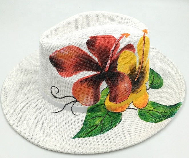 A white woven sun hat featuring a painted tropical hibiscus flower design in red, yellow, and green on the front panel and brim.